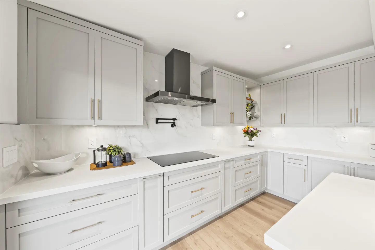 Bright kitchen with gray cabinets, white countertops, and stainless steel range hood. Wood flooring and marble backsplash.