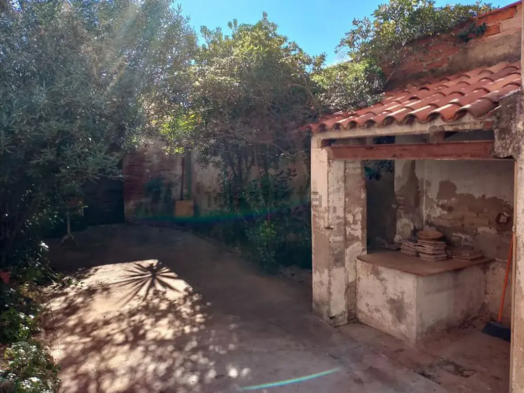 A rustic outdoor patio with a weathered stone structure, terracotta roof tiles, lush trees, and a sunlit concrete floor.