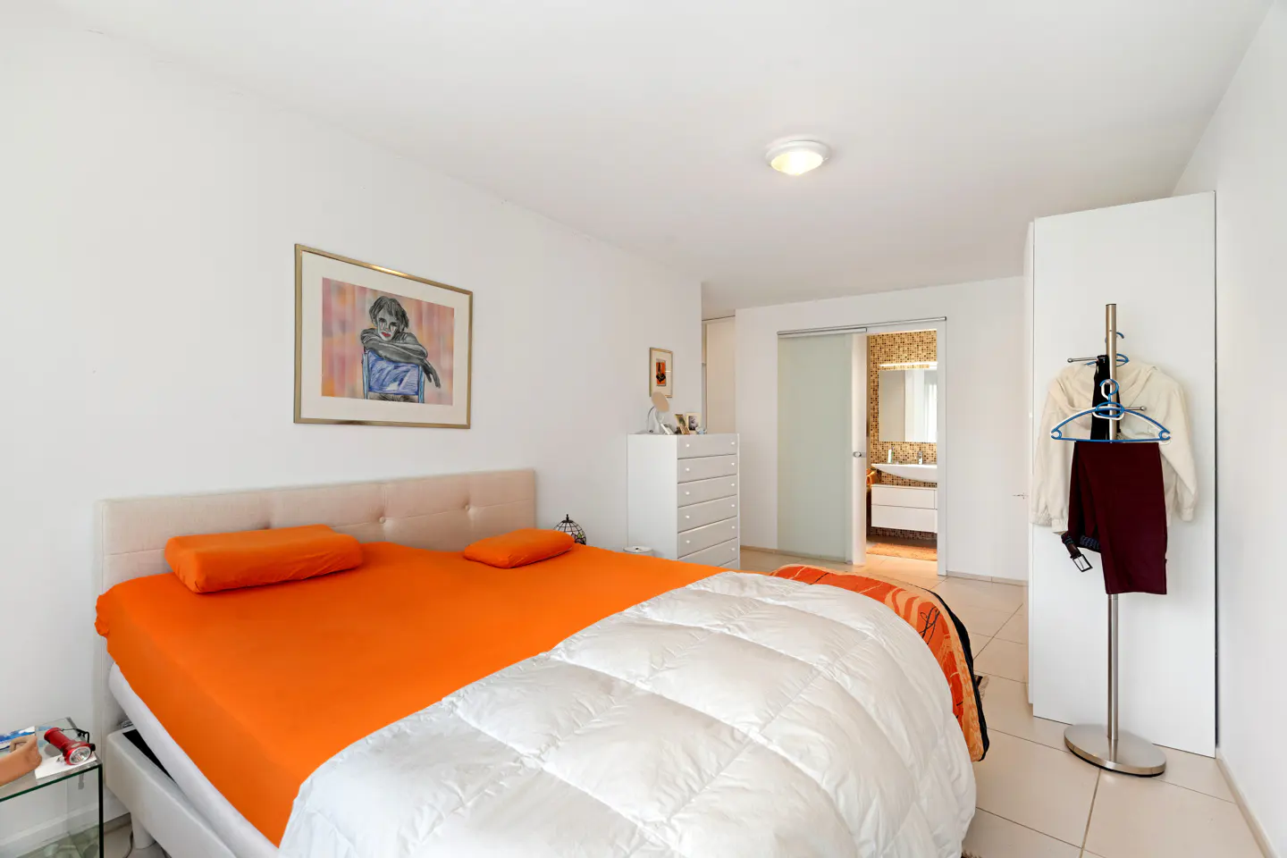 Bright bedroom with orange sheets, white duvet, and art. Bathroom visible through sliding glass door. Clothes hang on a rack.