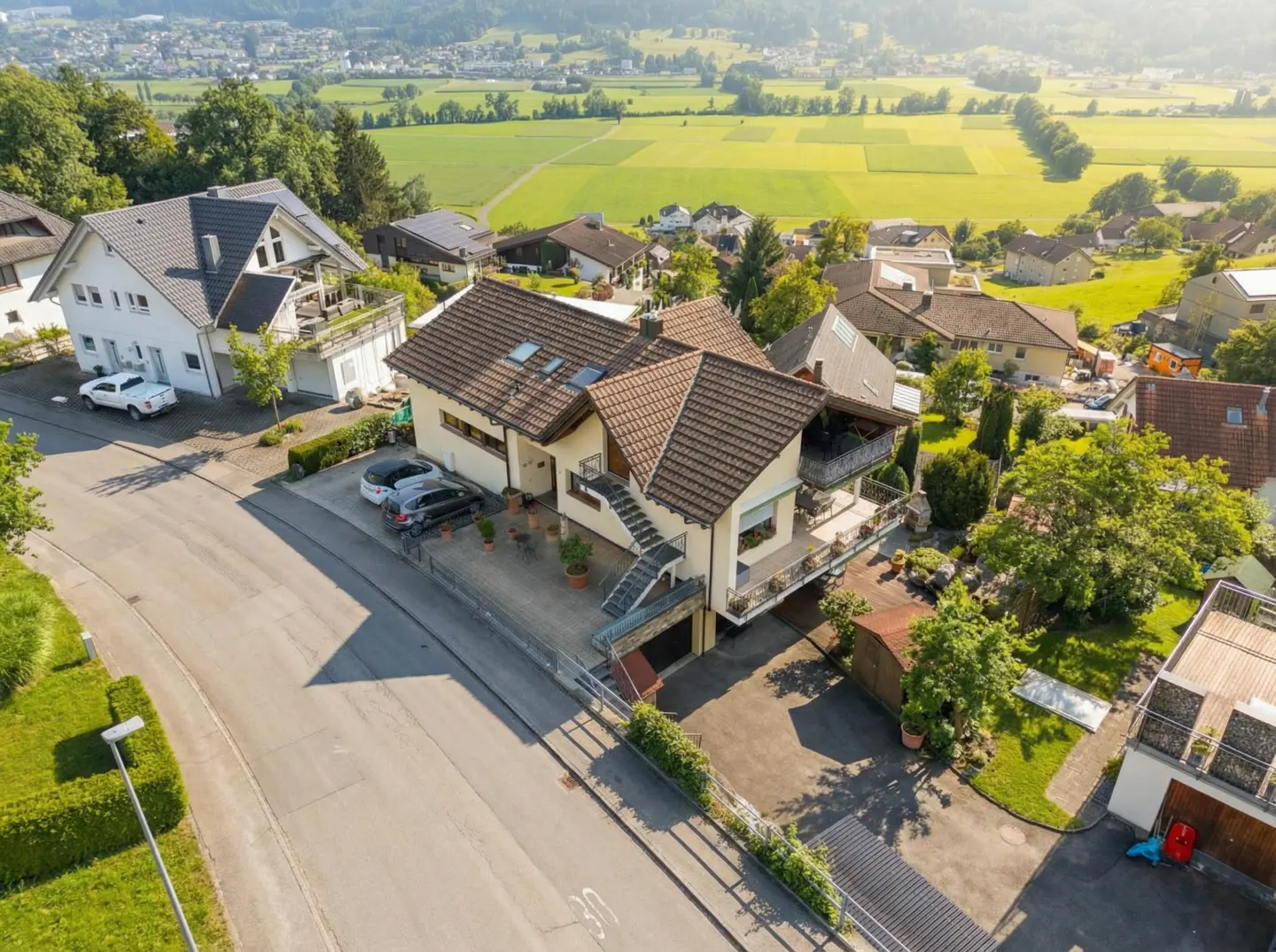 Aerial view of a beige two-story house with a brown roof, balconies, and a driveway with parked cars. Green fields and hills are in the background.
