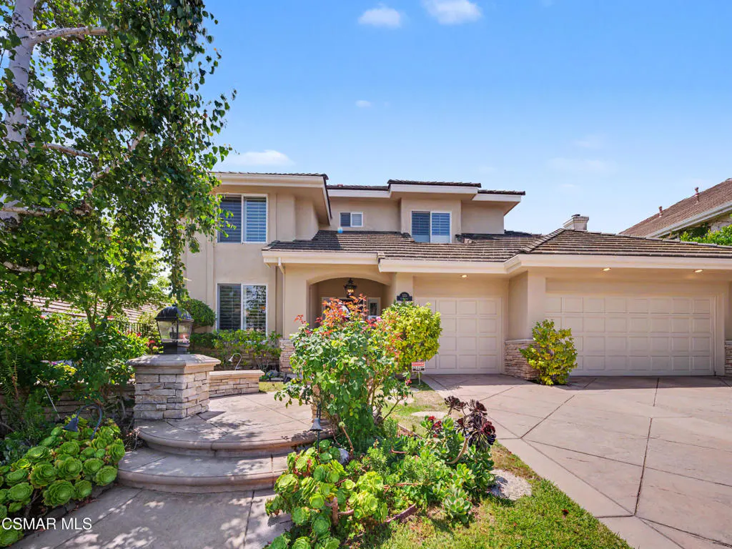 Two-story beige suburban home with a multi-car garage, stone-accented entryway, and lush front yard landscaping.