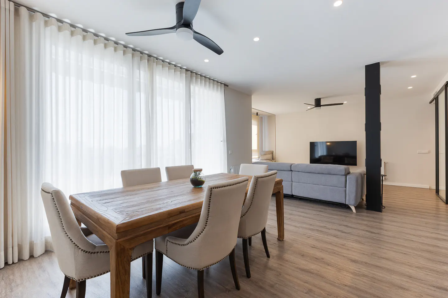 Bright, open-concept living space with a wood dining table, cream chairs, and a gray sofa. A black ceiling fan is visible.