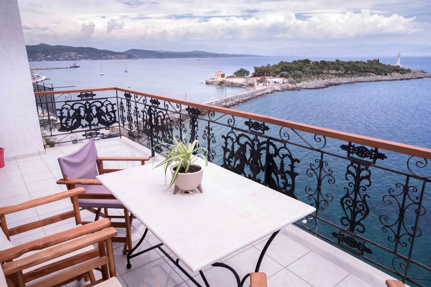 Balcony with ornate railing overlooks blue ocean and island with lighthouse. Table, chairs, and potted plant on tiled floor.