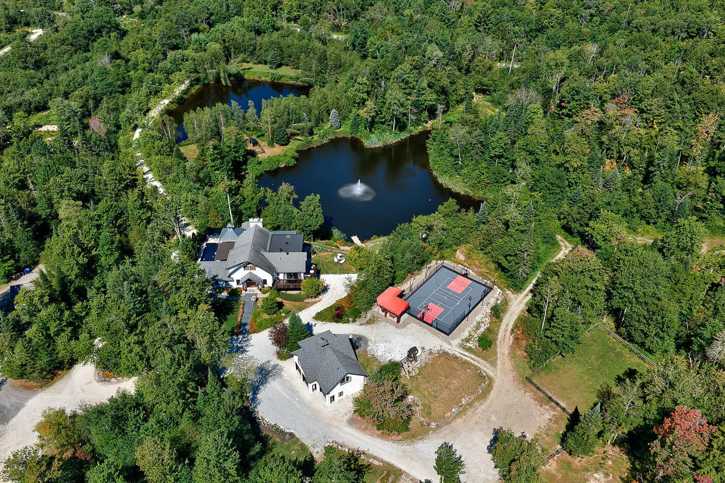 Aerial view of a property with a large pond, fountain, tennis court, and two houses surrounded by lush green trees.