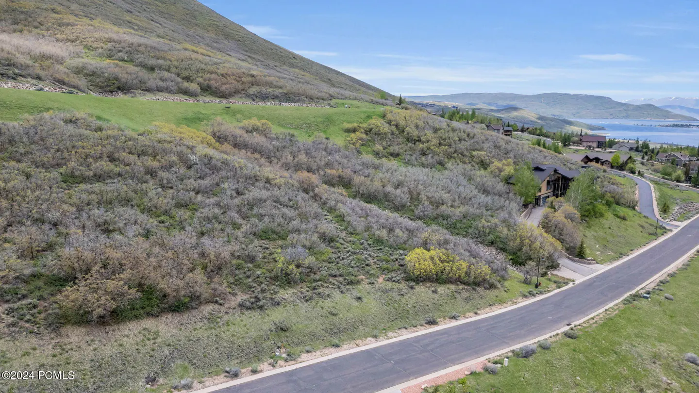 A hillside view shows a road leading to a house, with a lake and mountains in the background under a blue sky.