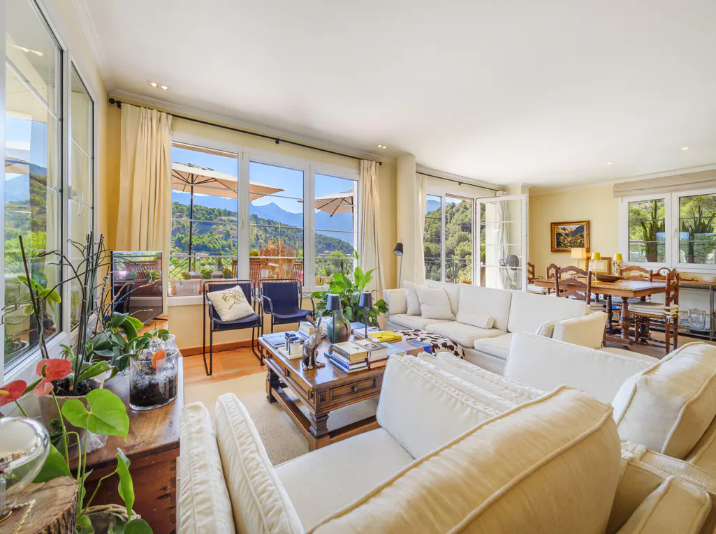 Bright living room with white sofas, wood table, and large windows showing mountain views. Plants add a touch of nature.