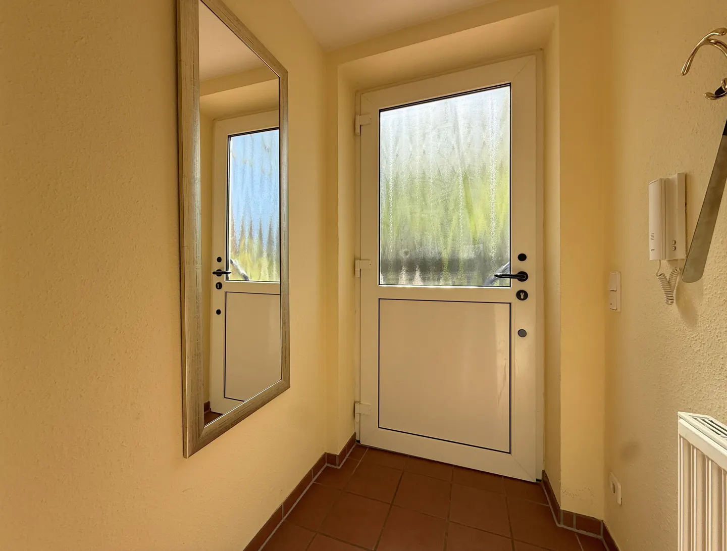 Entryway with a white door, a mirror reflecting the door, and yellow walls. A coat rack and intercom are on the right wall.