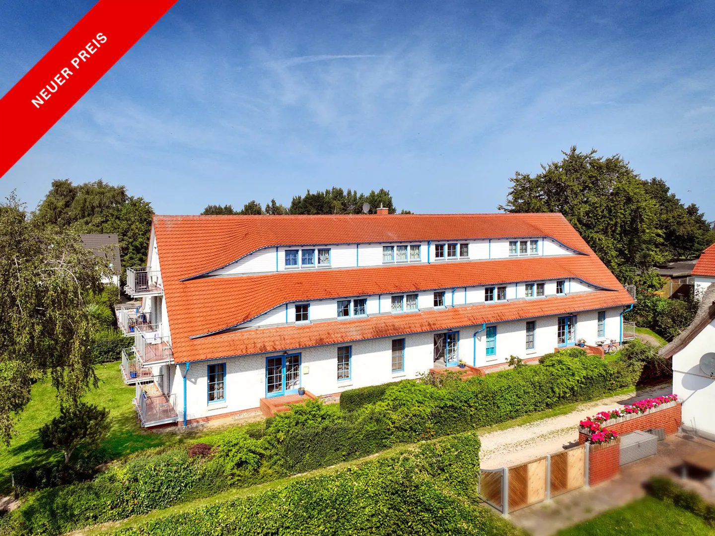 Two-story white building with a red tile roof and blue trim, surrounded by green trees and bushes. A red banner in the corner reads "Neuer Preis".