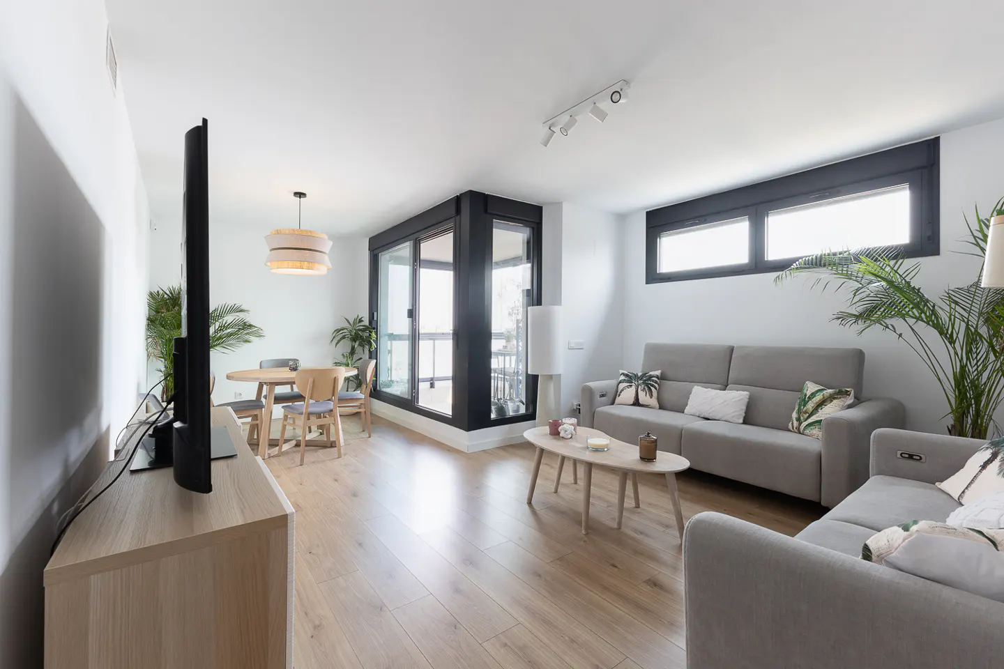 Bright living room with wood floors, gray sofas, a wood table, and a TV on a light wood stand. Large windows let in natural light.