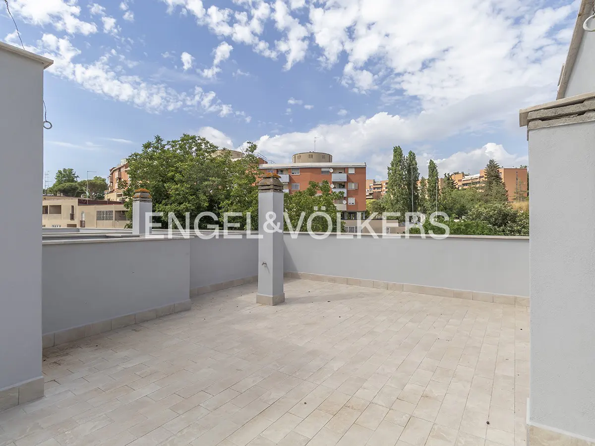 Wide rooftop terrace with gray walls and light tile flooring, overlooking trees and buildings under a cloudy sky.