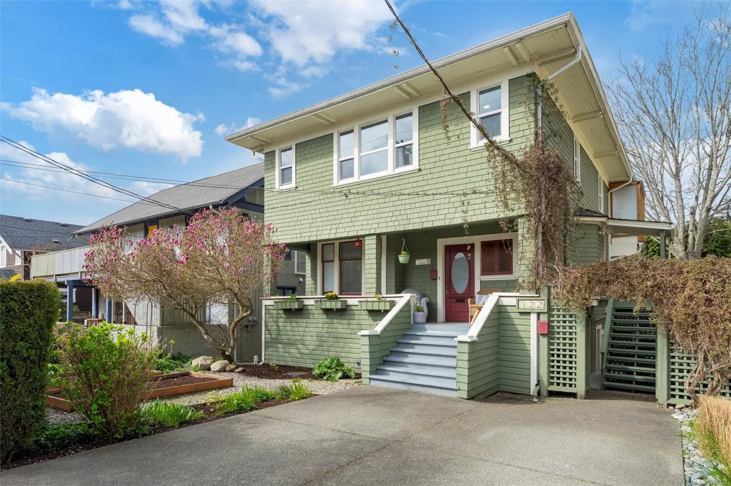 Two-story green house with a red door and white trim, gray stairs, and a concrete driveway. A tree with pink flowers is in the front yard.