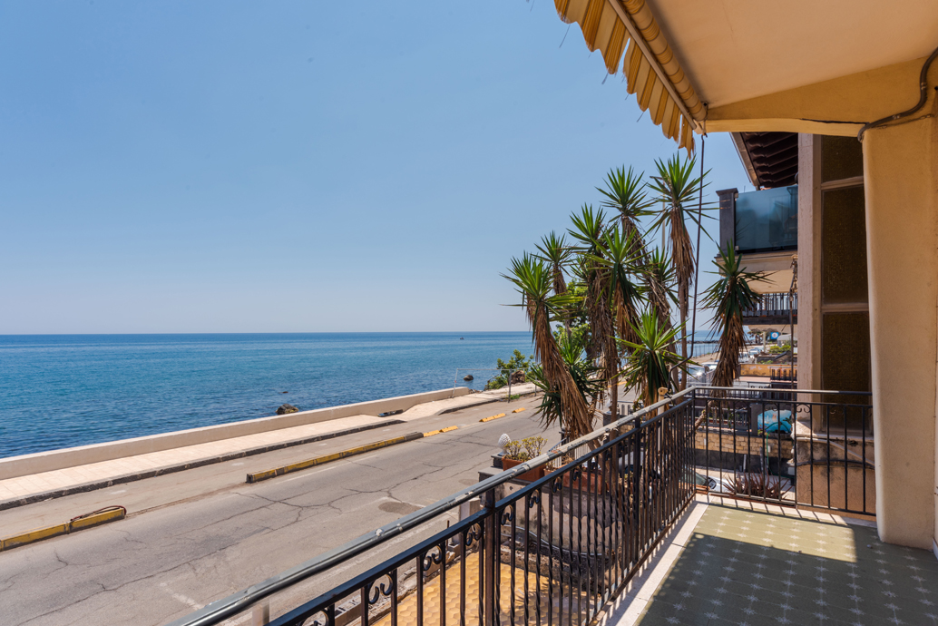 Balcony view overlooking a coastal road and the blue sea, featuring palm trees and a metal railing under a yellow awning.
