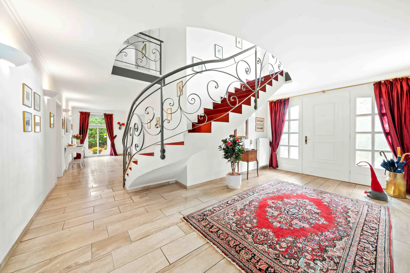 Bright foyer with a curved staircase, red accents, and a patterned rug. White walls and doors, with red curtains framing the entrance.