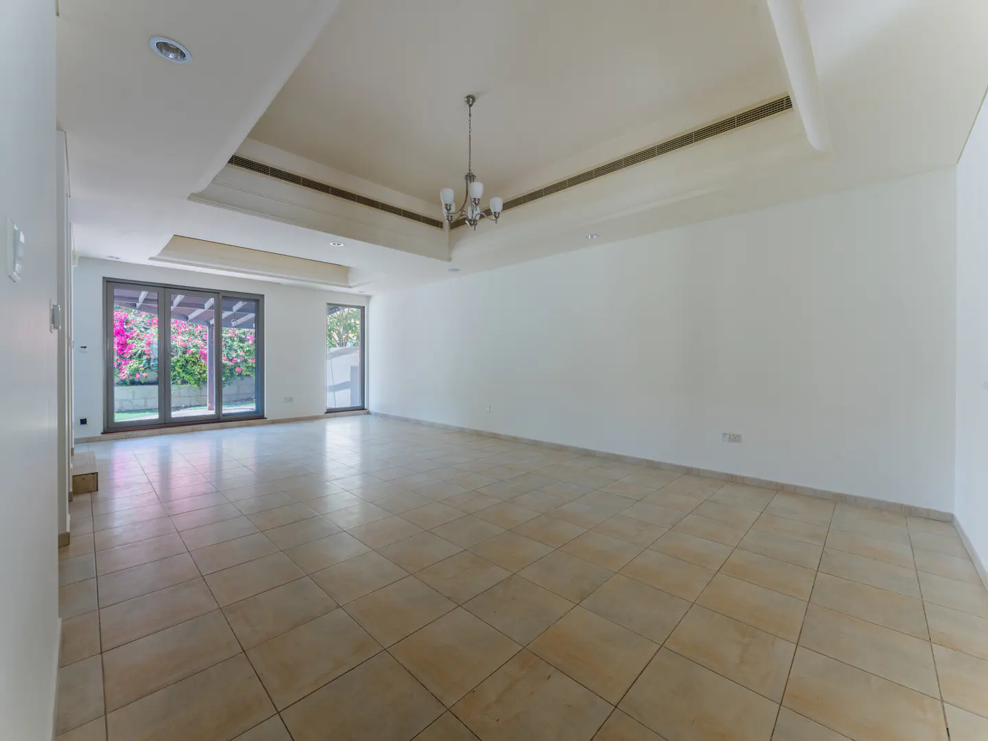A bright, empty room with beige tile floors, white walls, and a chandelier. Sliding glass doors lead to a garden with pink flowers.