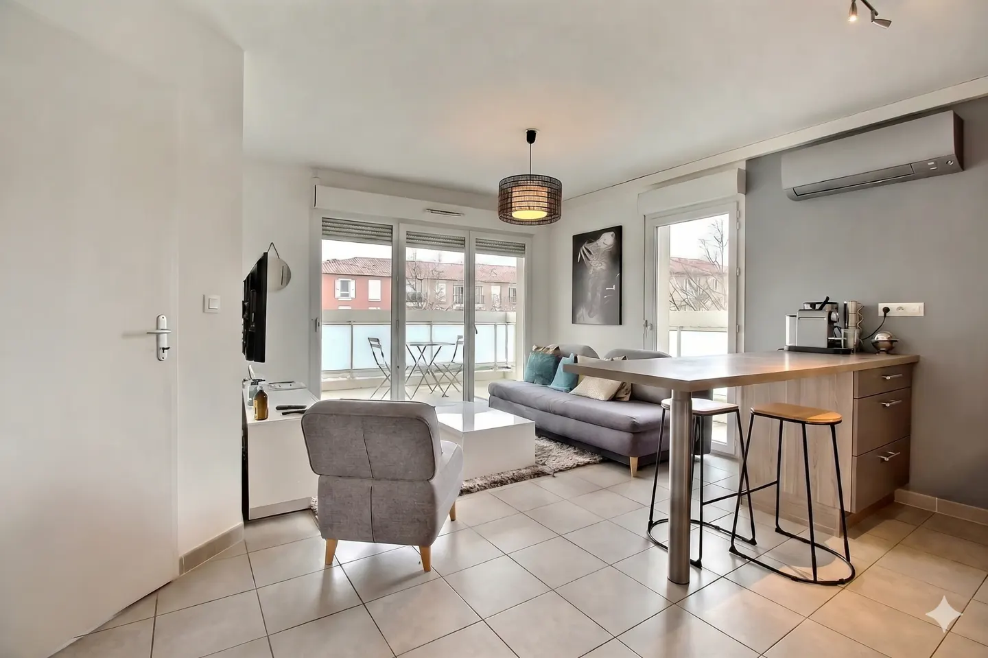 Bright apartment interior with gray sofa, chair, and island with stools. Balcony with table and chairs visible through sliding glass doors.