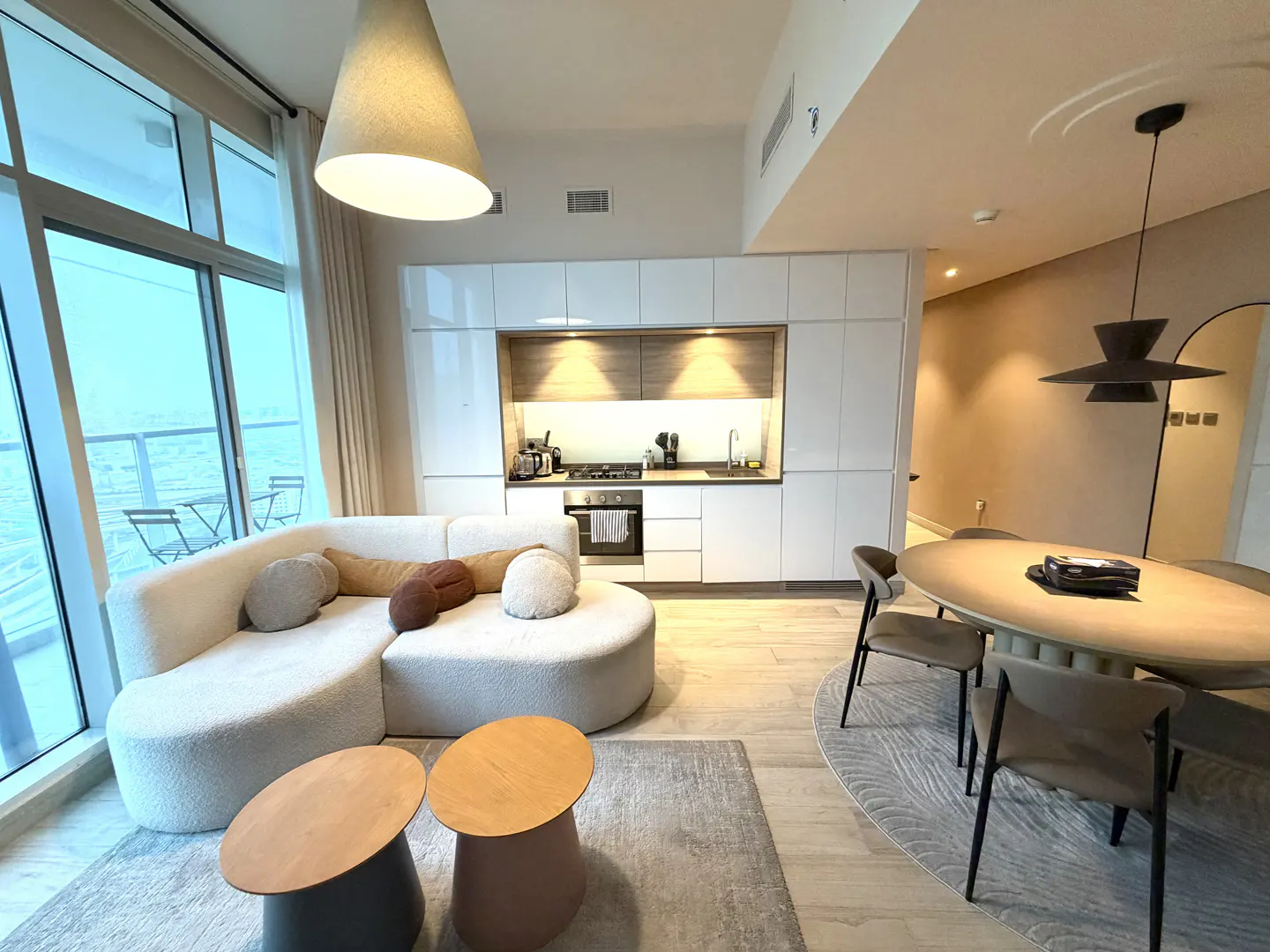 A modern apartment interior with a white sofa, kitchen, and dining area. Large windows offer a view of the ocean.