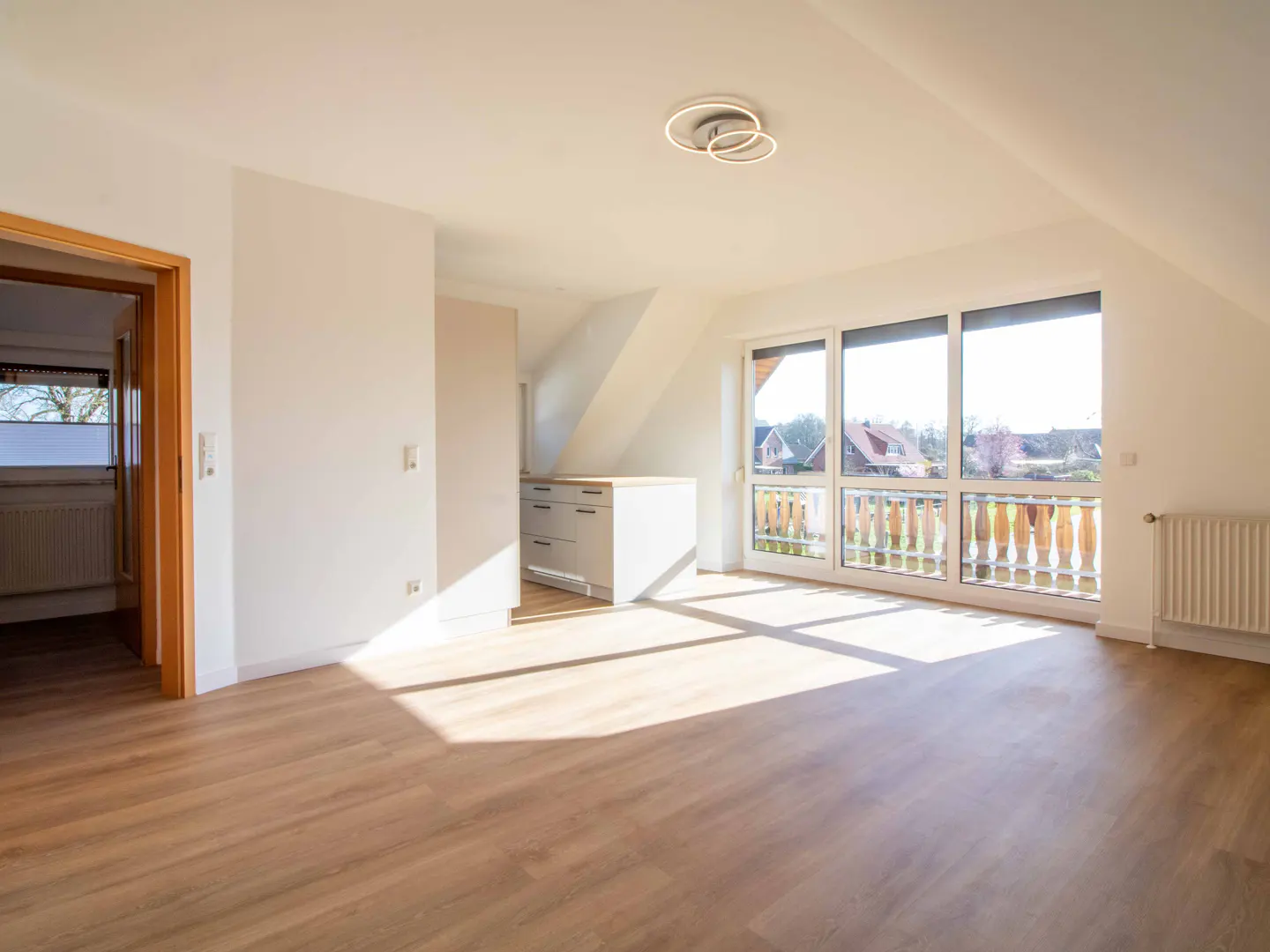 Bright, empty attic apartment with wood floors, white walls, and large windows overlooking a neighborhood.