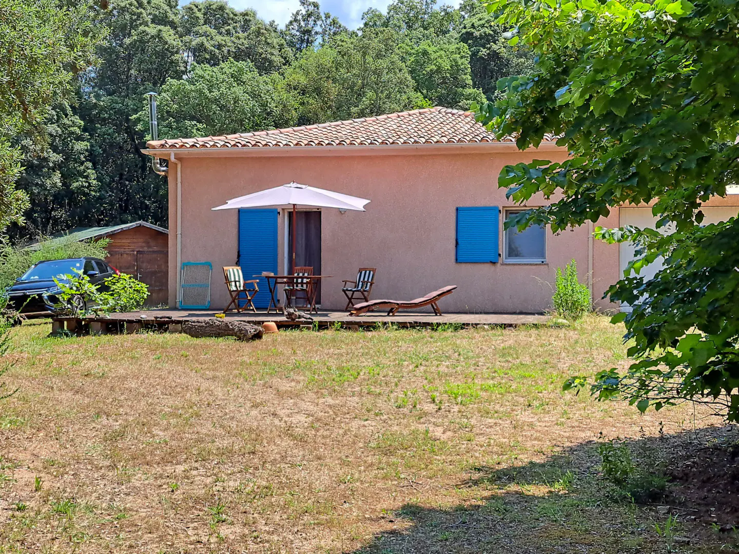 A single-story house with a tan exterior, blue shutters, and a tiled roof. Outdoor furniture sits on a patio, with a lawn in front and trees in the background.