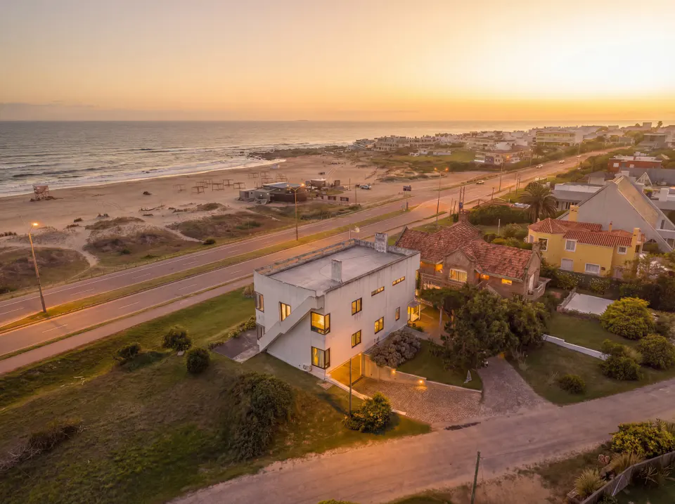 Aerial view of a modern white house near a beach at sunset, with a road, other houses, and the ocean visible.
