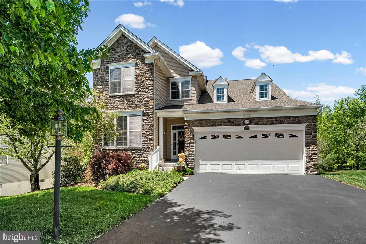 Two-story house with stone and beige siding, white garage door, and black asphalt driveway. Green lawn and trees surround the house.
