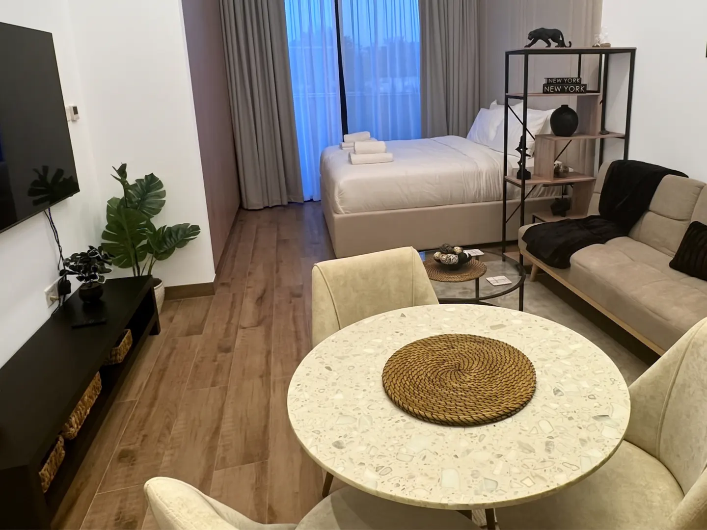 A modern studio apartment with a round table, chairs, sofa, bed, TV, and shelving unit. The color palette is neutral with beige, brown, and black accents.