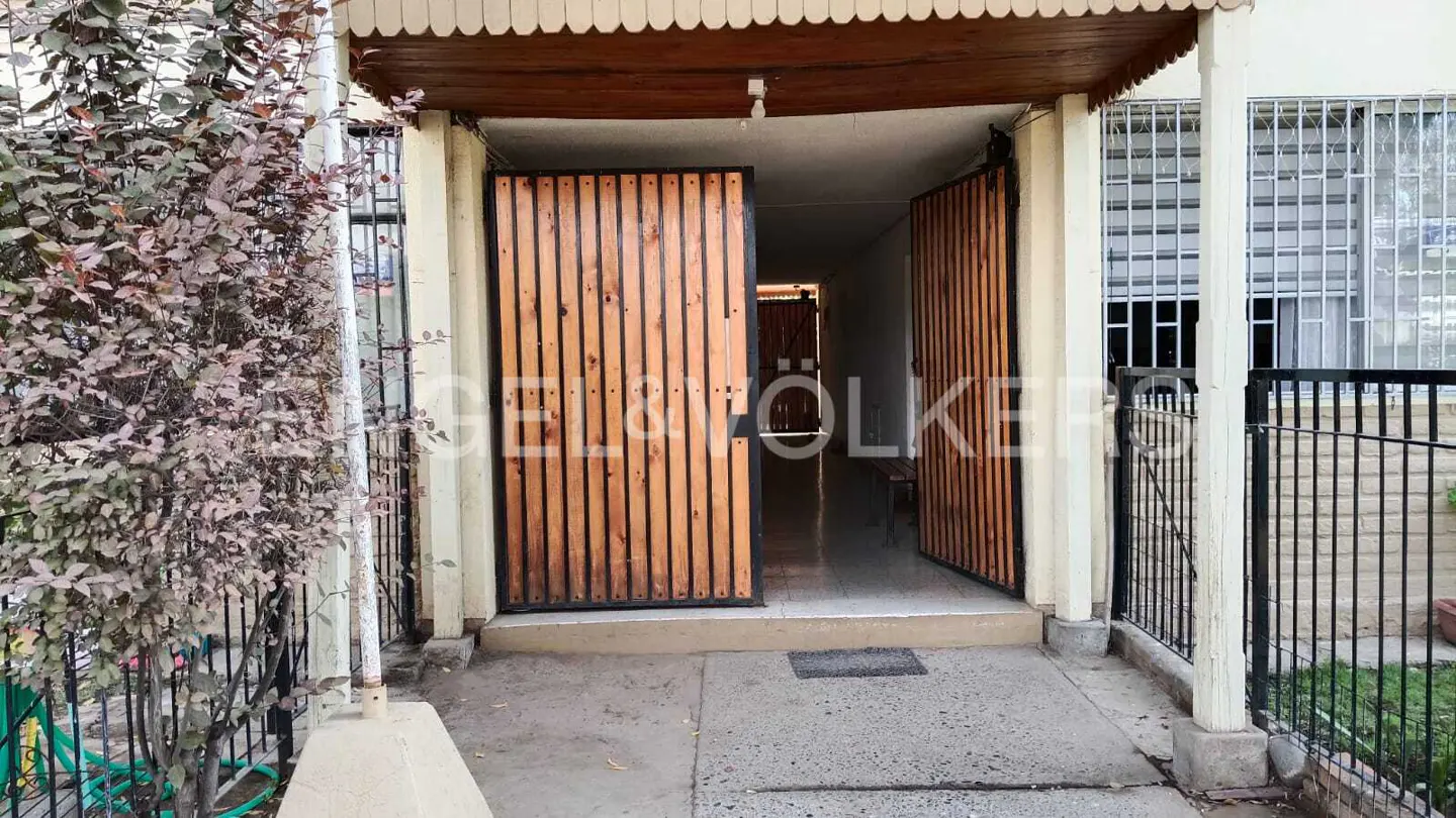 Entrance to a building with open, light-wood doors and a concrete walkway. A tree is on the left, and a black fence is on the right.