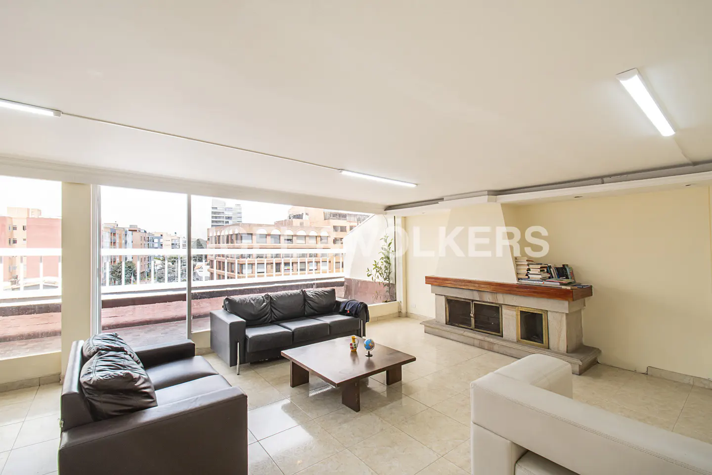 Bright living room with black leather sofas, a wooden coffee table, and a stone fireplace. Large windows offer city views.