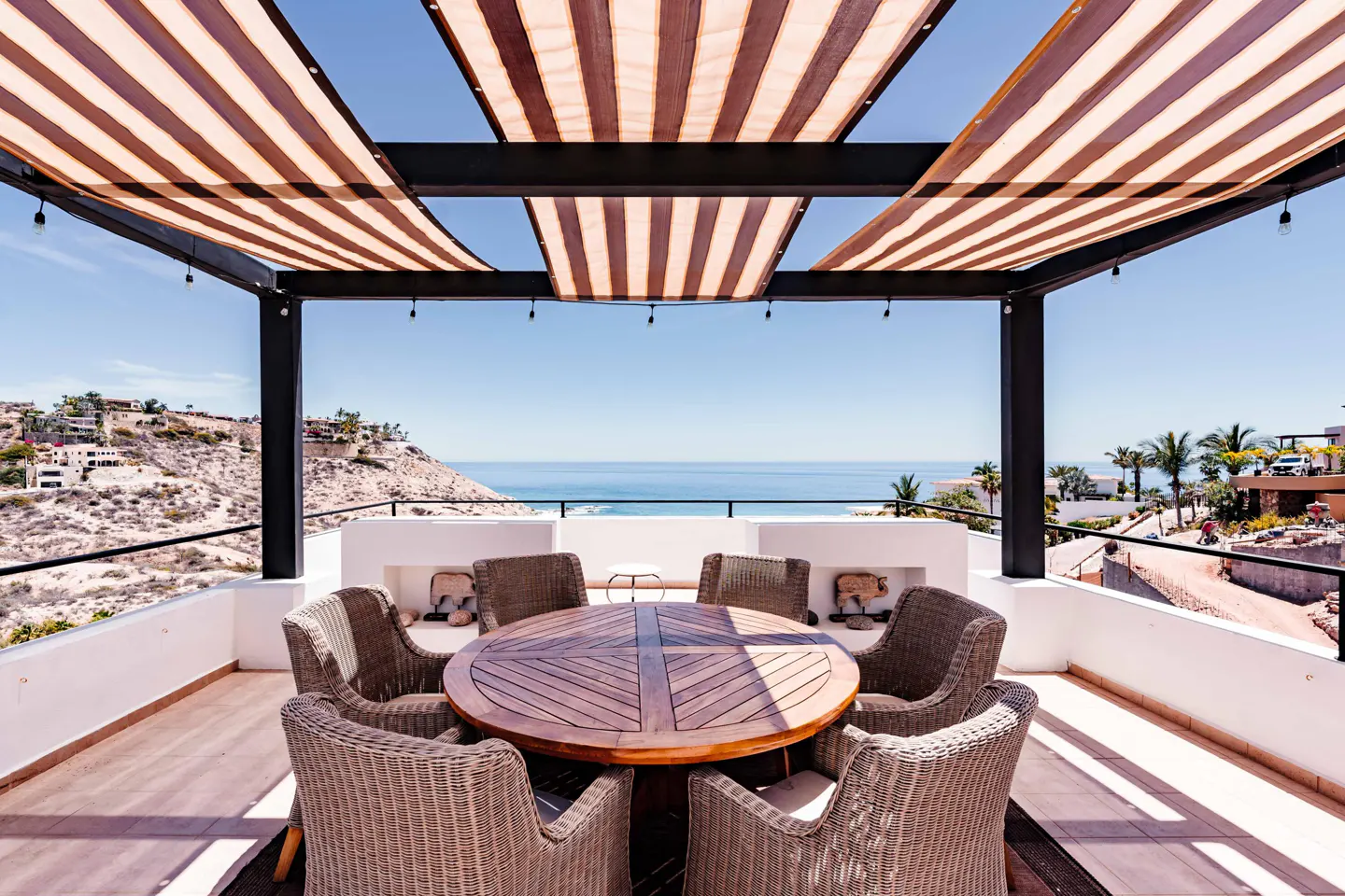 Outdoor patio with a round wooden table and wicker chairs under a striped awning, overlooking the ocean.