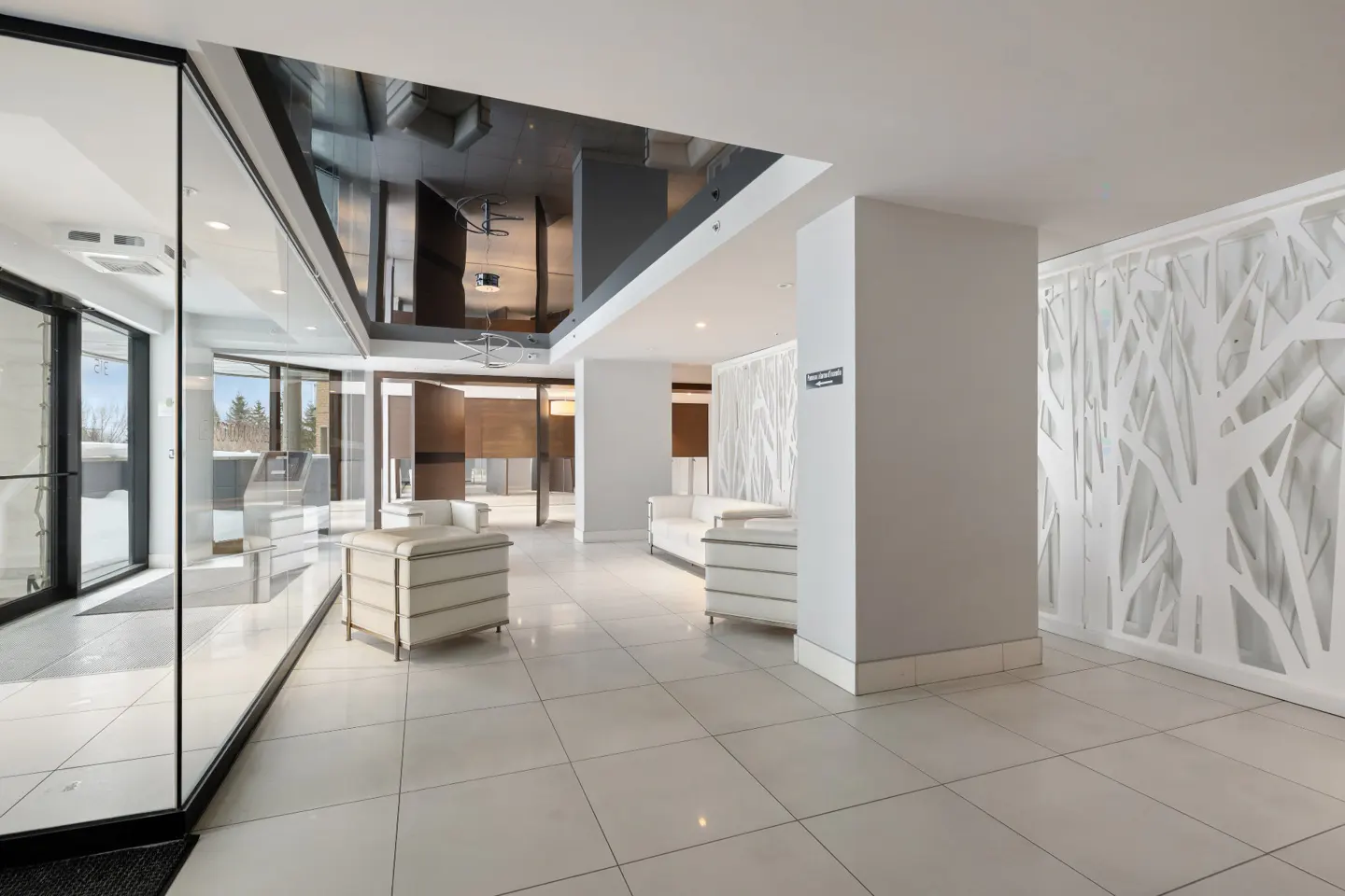 Modern lobby with white tile floors, white couches, and a wall with a white tree branch design. Glass doors lead outside.
