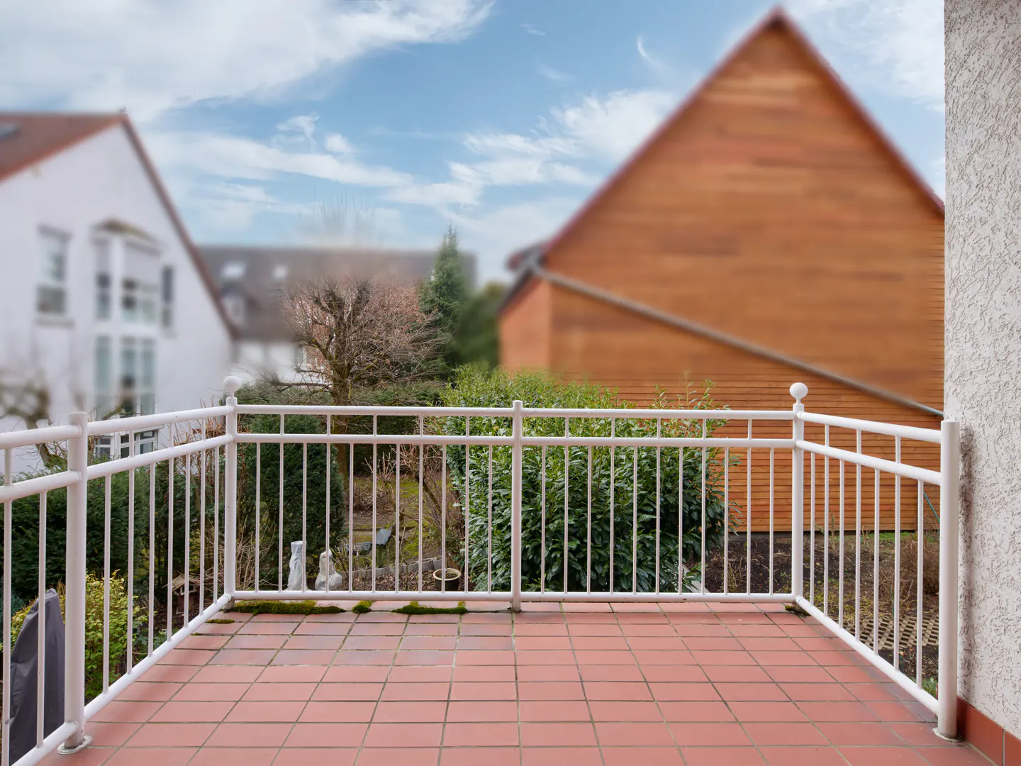 Balcony with red tile floor and white railing overlooking a garden and houses under a blue sky.