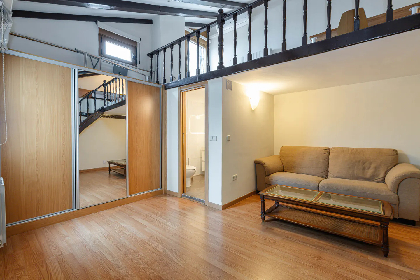 Living room with wood floors, a beige sofa, and a glass-topped coffee table. A loft with a dark railing overlooks the space.