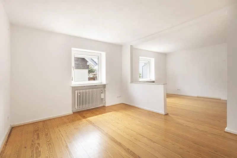 Bright, empty room with hardwood floors, white walls, and two windows. A radiator sits beneath one window. A partial wall divides the space.