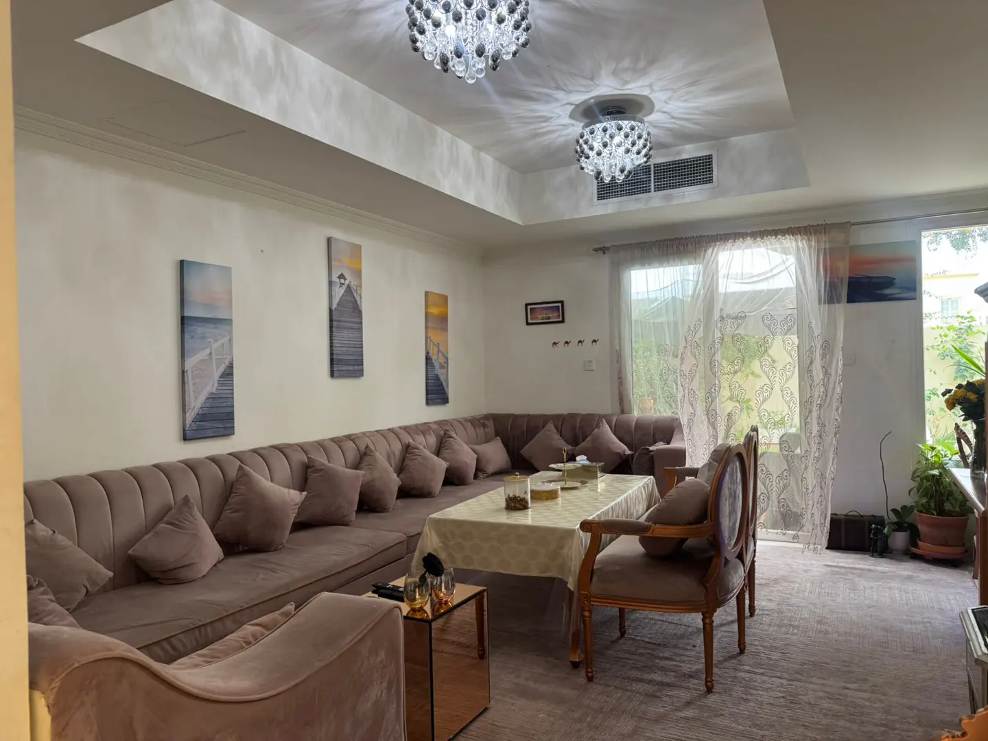 A living room with a long brown sofa, a table, and two chairs. There are paintings on the wall and two chandeliers on the ceiling.