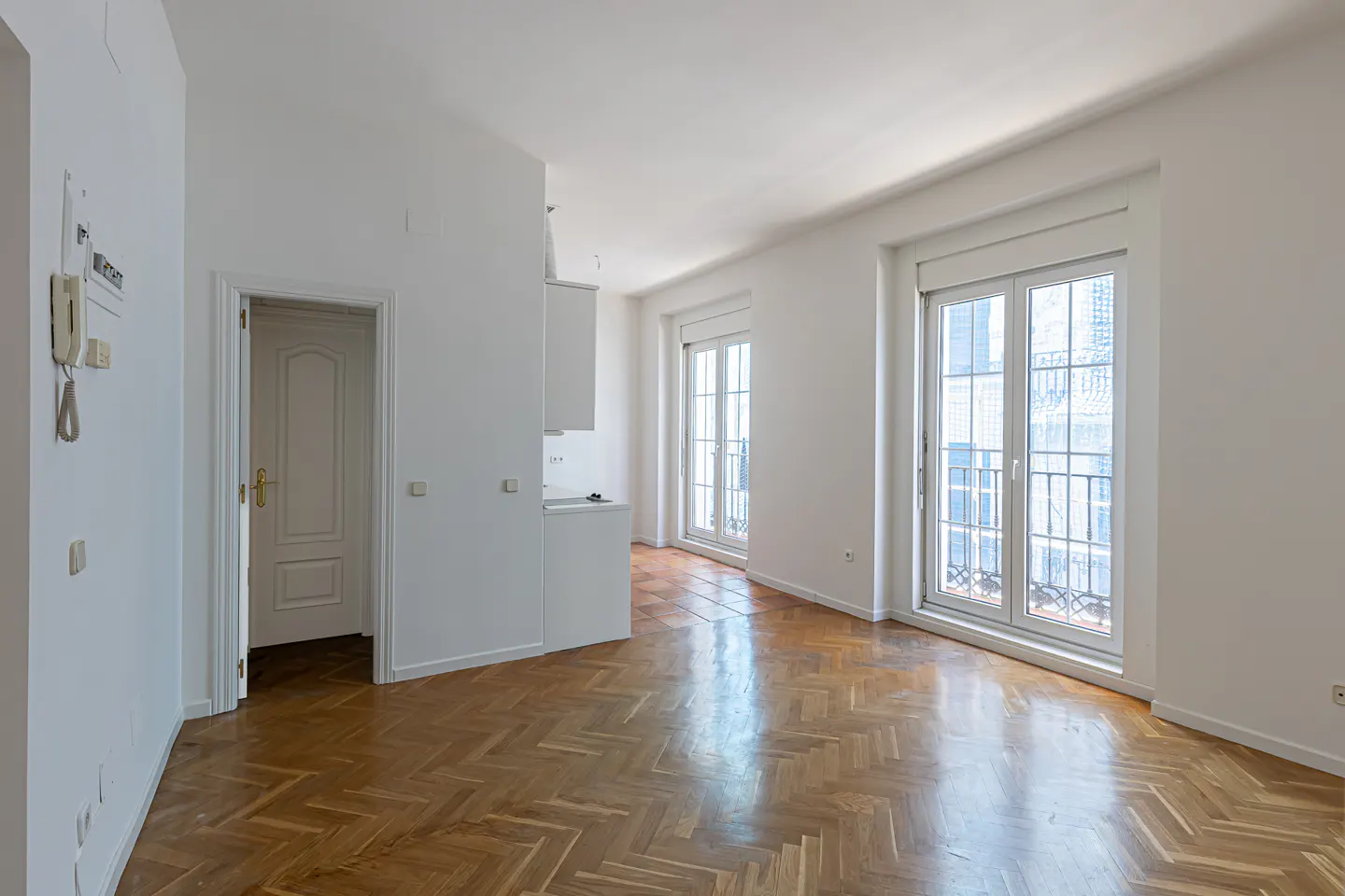 Bright, empty apartment with herringbone wood floors, white walls, and two sets of French doors leading to balconies.