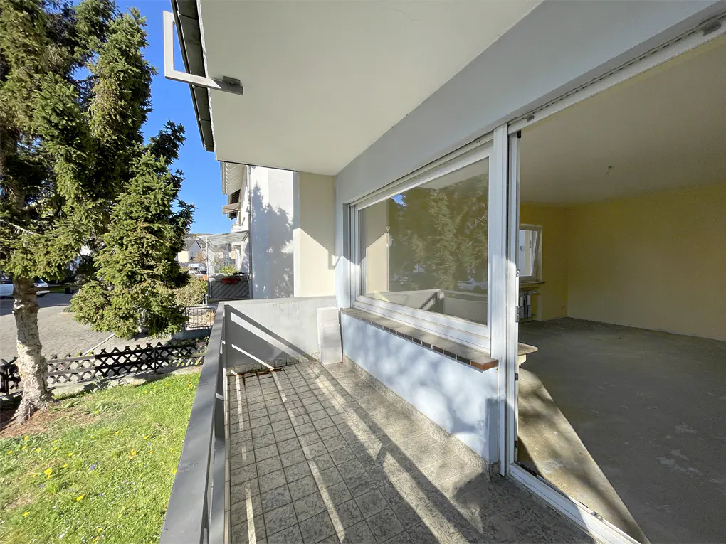 Balcony view with gray tile floor, metal railing, and open sliding glass door to a yellow room. Trees and grass visible outside.