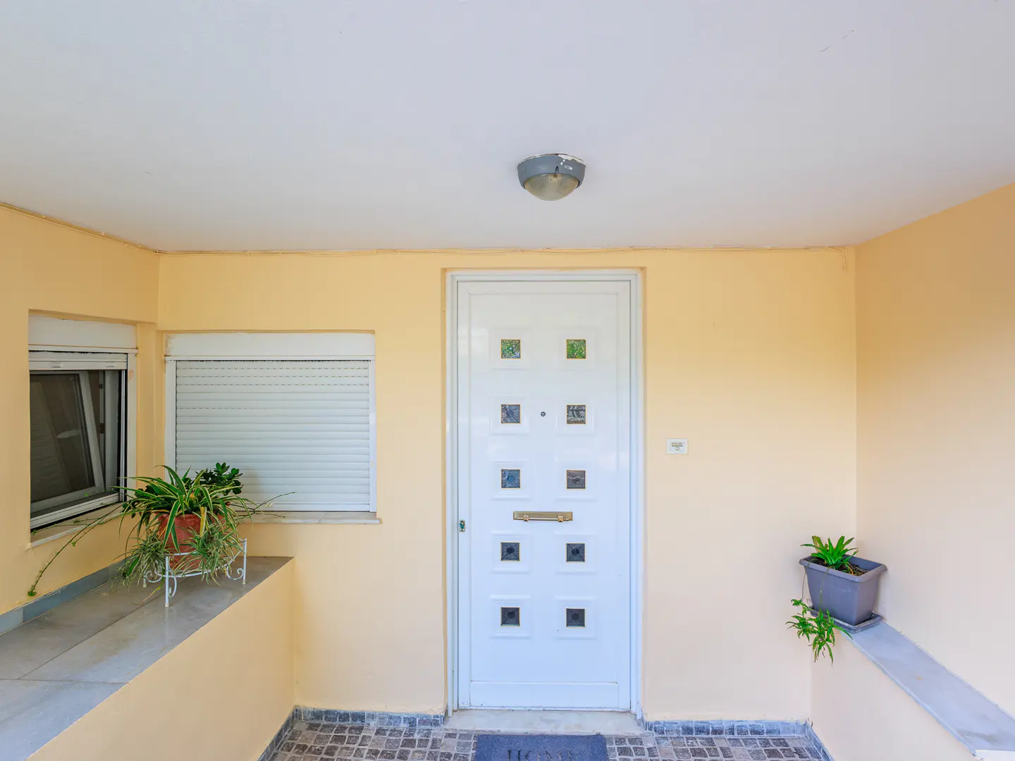 Covered porch with a white door, two windows, and potted plants. The walls are light yellow.