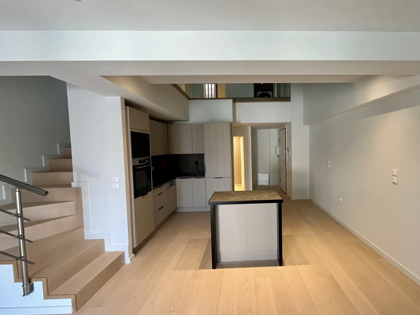 Open-concept kitchen and living area with light wood floors, white walls, and a staircase on the left. Kitchen island in the center.