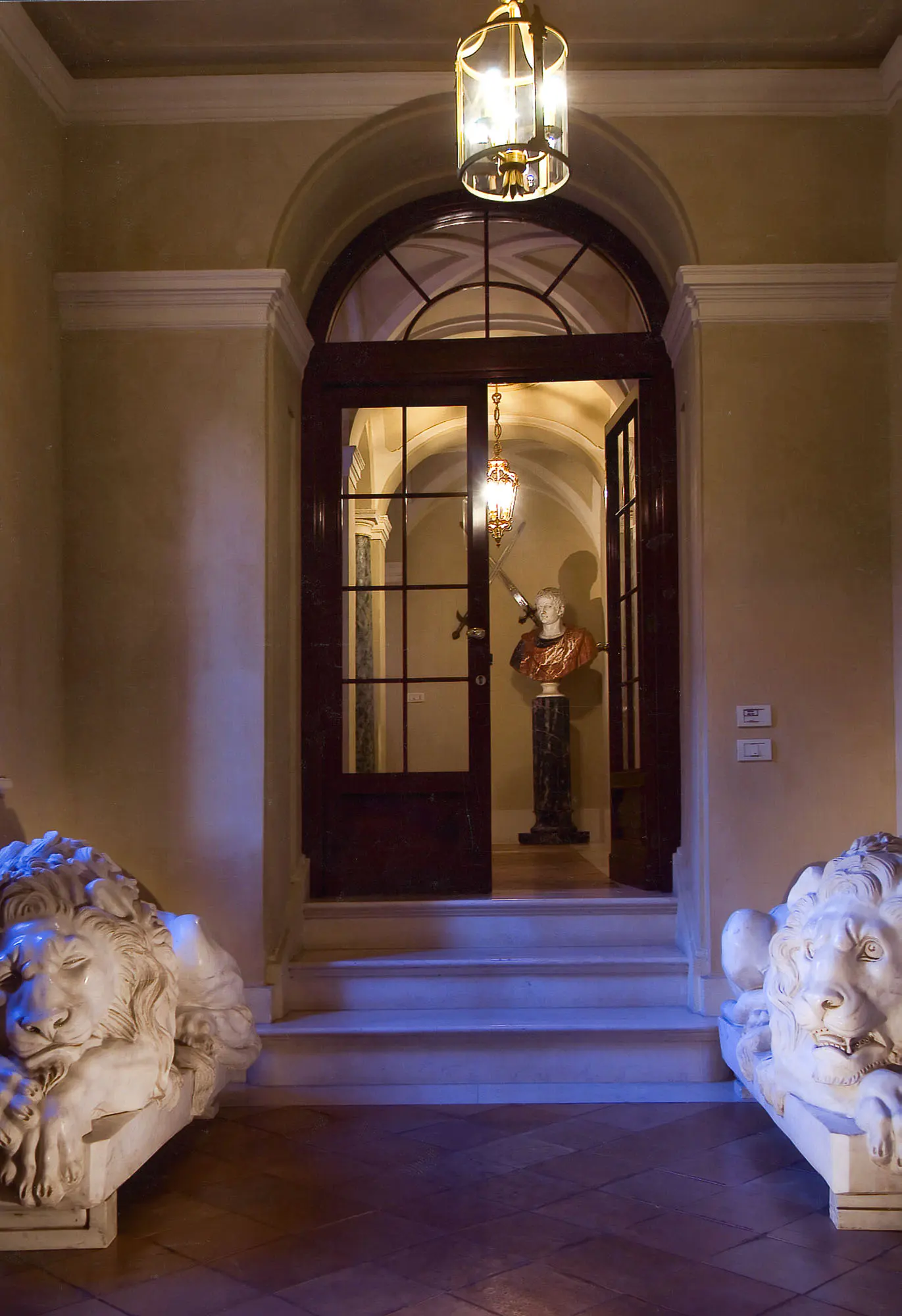 Interior view of a hallway with marble lion statues flanking steps leading to an open doorway with a bust statue inside.