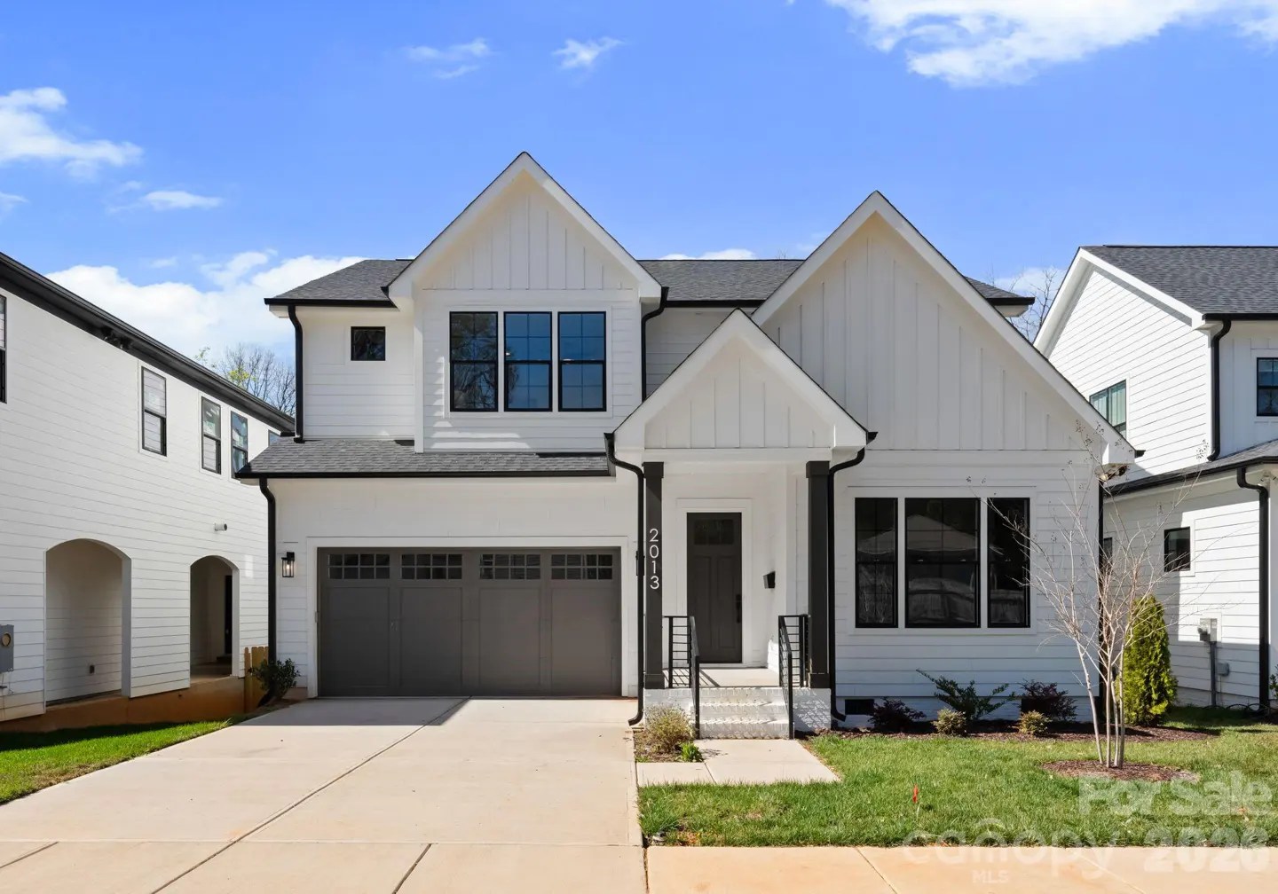 Modern two-story white farmhouse with a gray garage, black trim, and a concrete driveway under a blue sky.