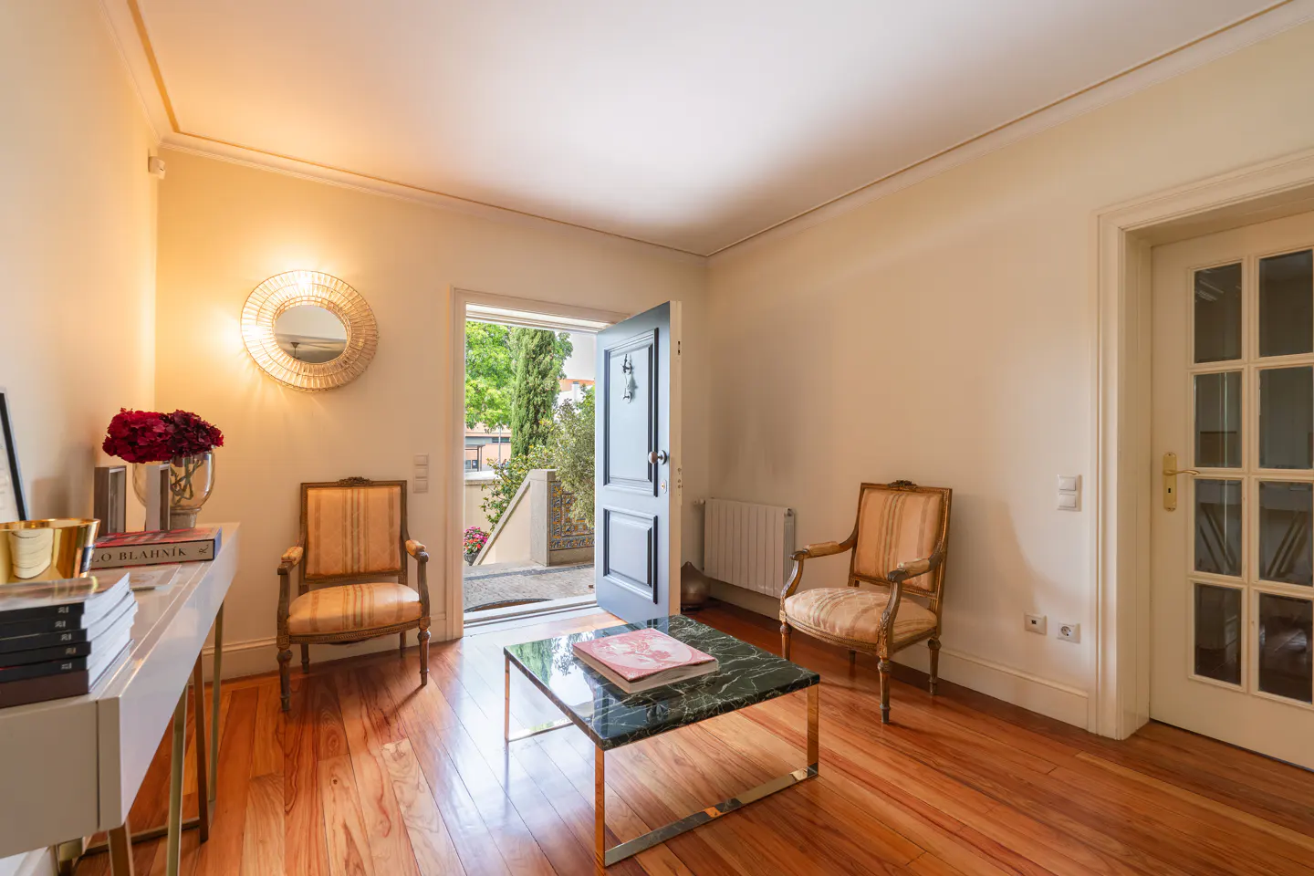 Living room with hardwood floors, two striped chairs, and a marble-topped coffee table. An open door leads to a patio with greenery.