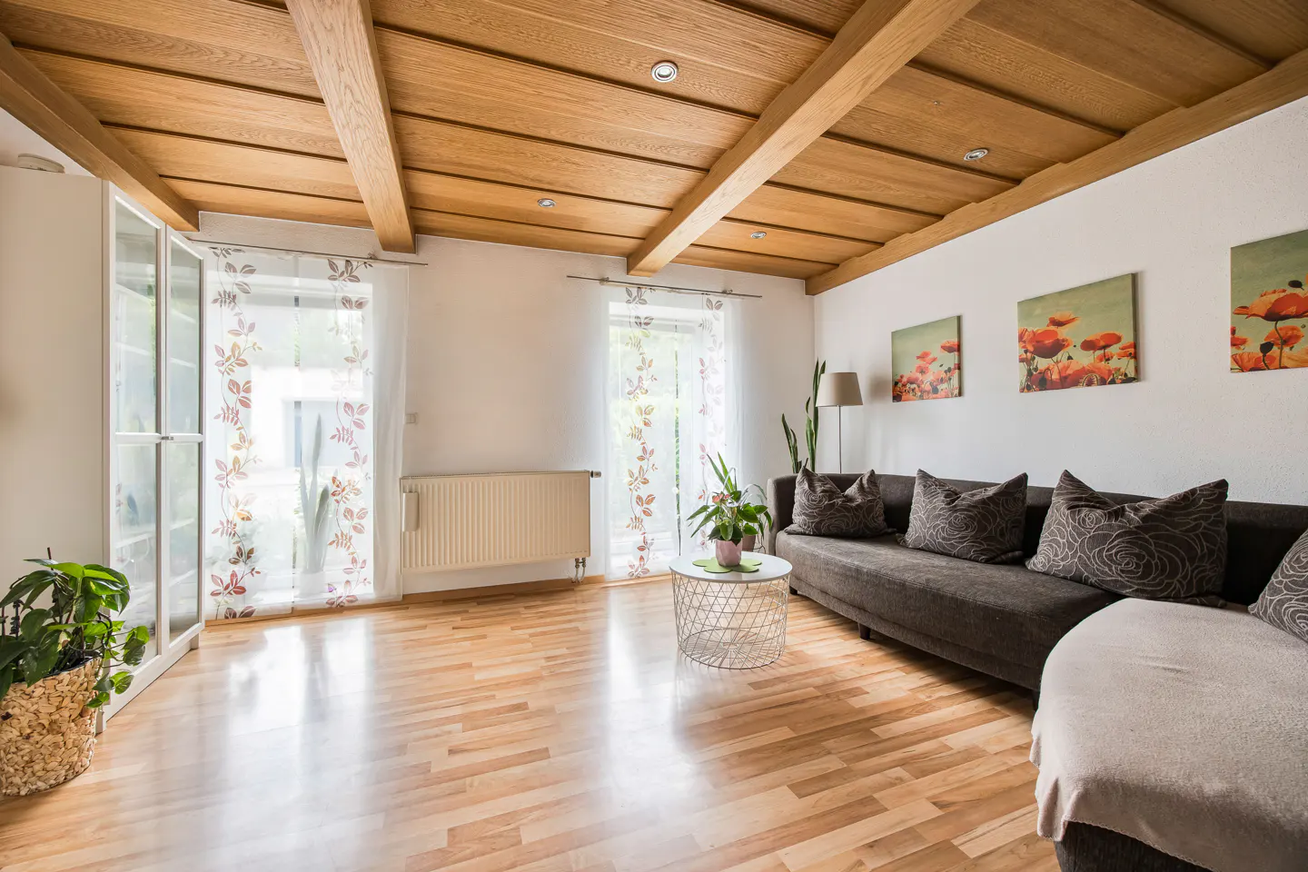 Bright living room with wood floors, beamed ceiling, and white walls. A gray sofa with floral pillows sits beneath poppy artwork. Floral curtains adorn windows.