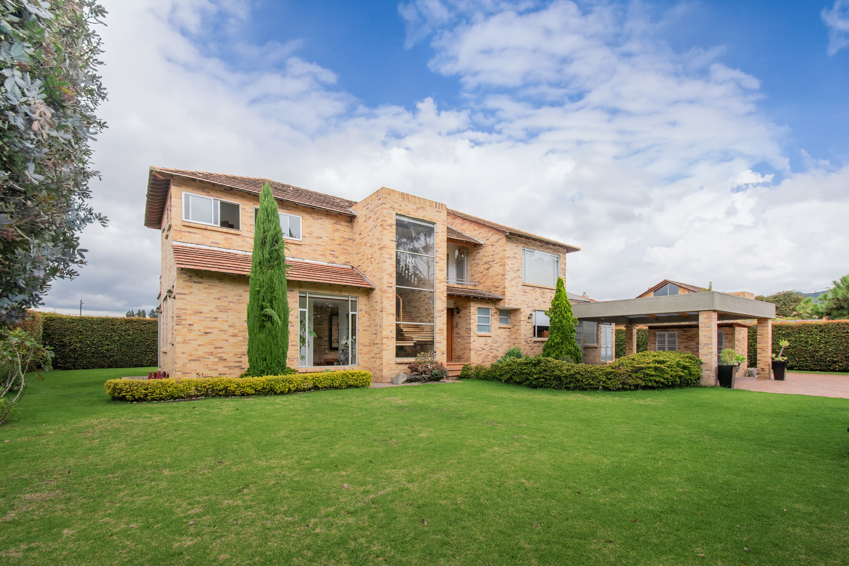 Two-story tan brick house with a large lawn, tall cypress tree, and attached carport under a blue sky with white clouds. [AI-generated text]