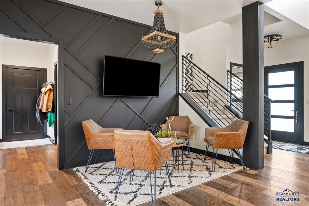 Modern living area with a black geometric accent wall, wall-mounted TV, three tan leather chairs, and a patterned rug.