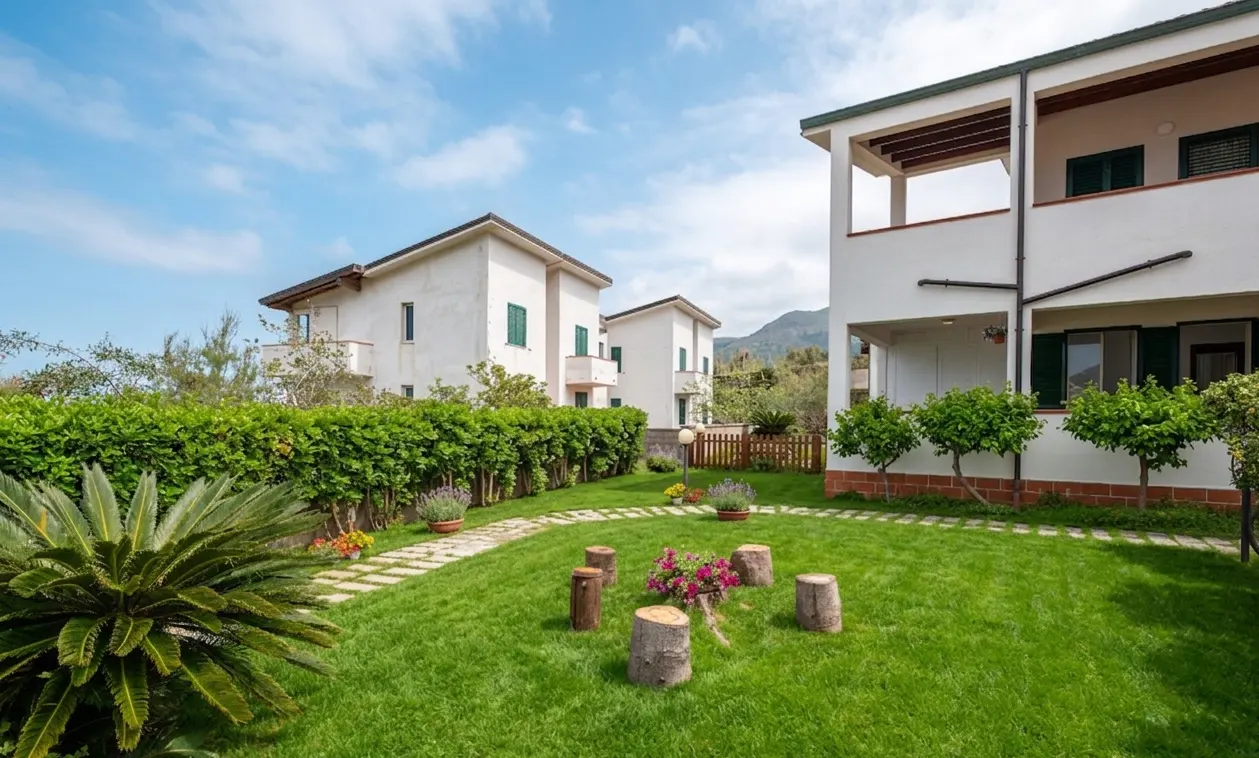 Exterior view of white houses with green shutters, a green lawn, and tree stump seating.