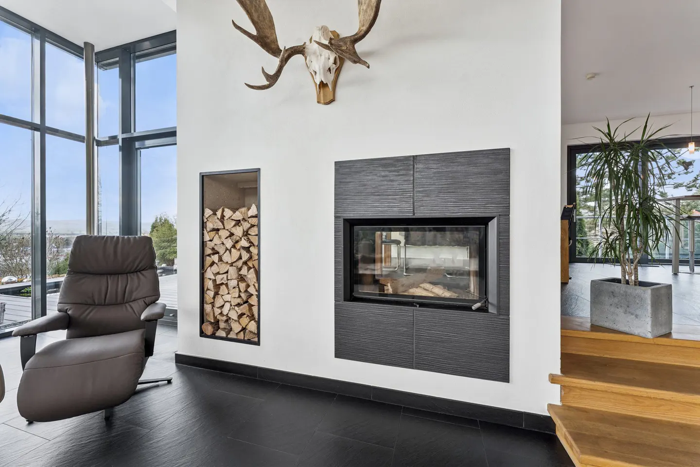 Living room with a black fireplace, firewood storage, and moose antlers above. A brown leather chair sits near a large window.