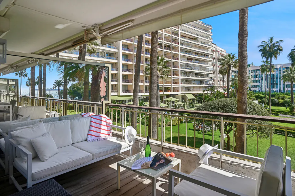 Balcony view with white furniture, a glass table with a magazine, and a view of palm trees, a lawn, and a building.