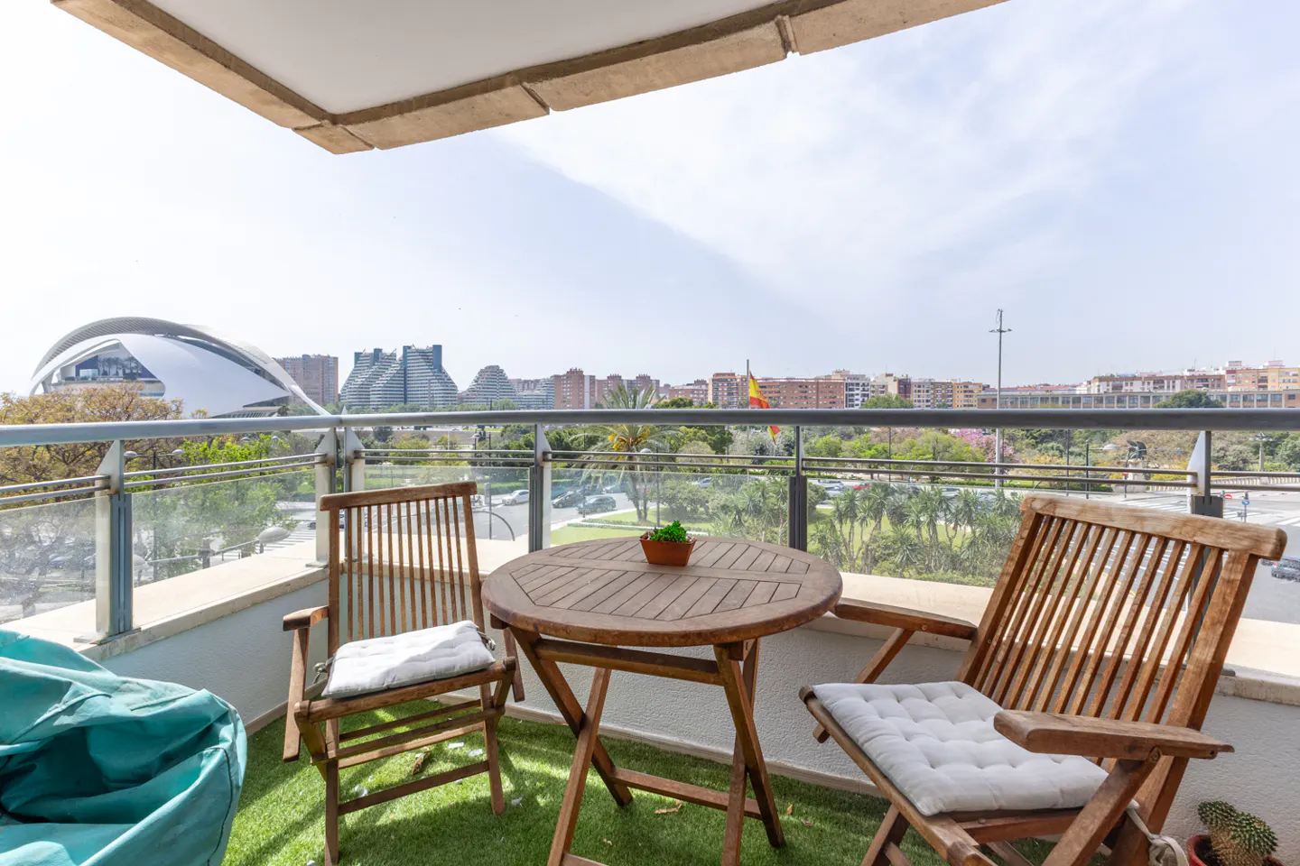Balcony with wooden table, chairs, and beanbag. City view with modern architecture and green space in the background.