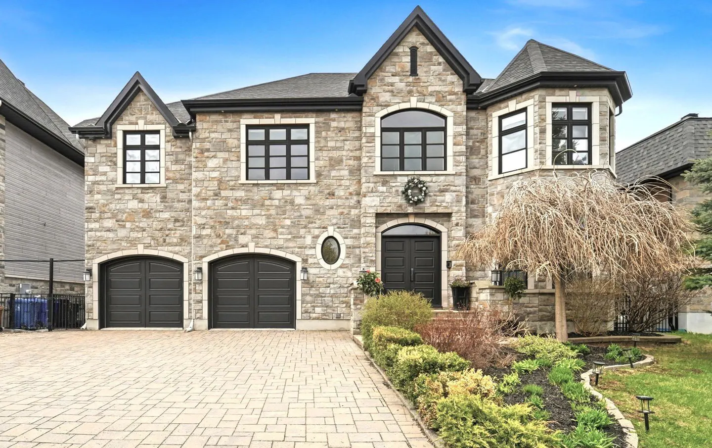 Two-story stone house with a double garage, black trim, arched entryway, and a paved driveway under a clear blue sky.