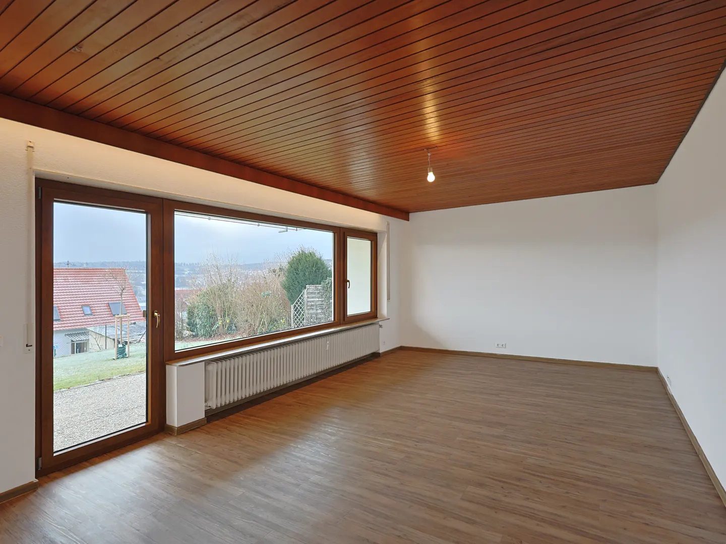 A bright, empty room with wood floors, a wood-paneled ceiling, and a large window overlooking a residential area.