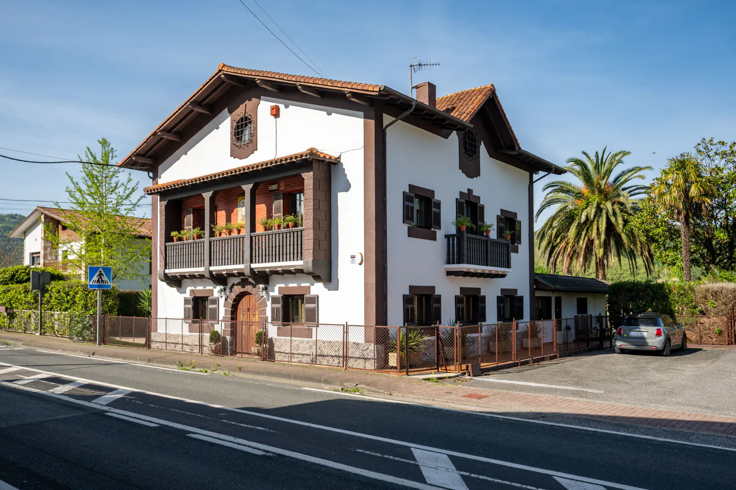 Two-story white house with brown trim, balconies with potted plants, and a red tile roof. A car is parked in the driveway.